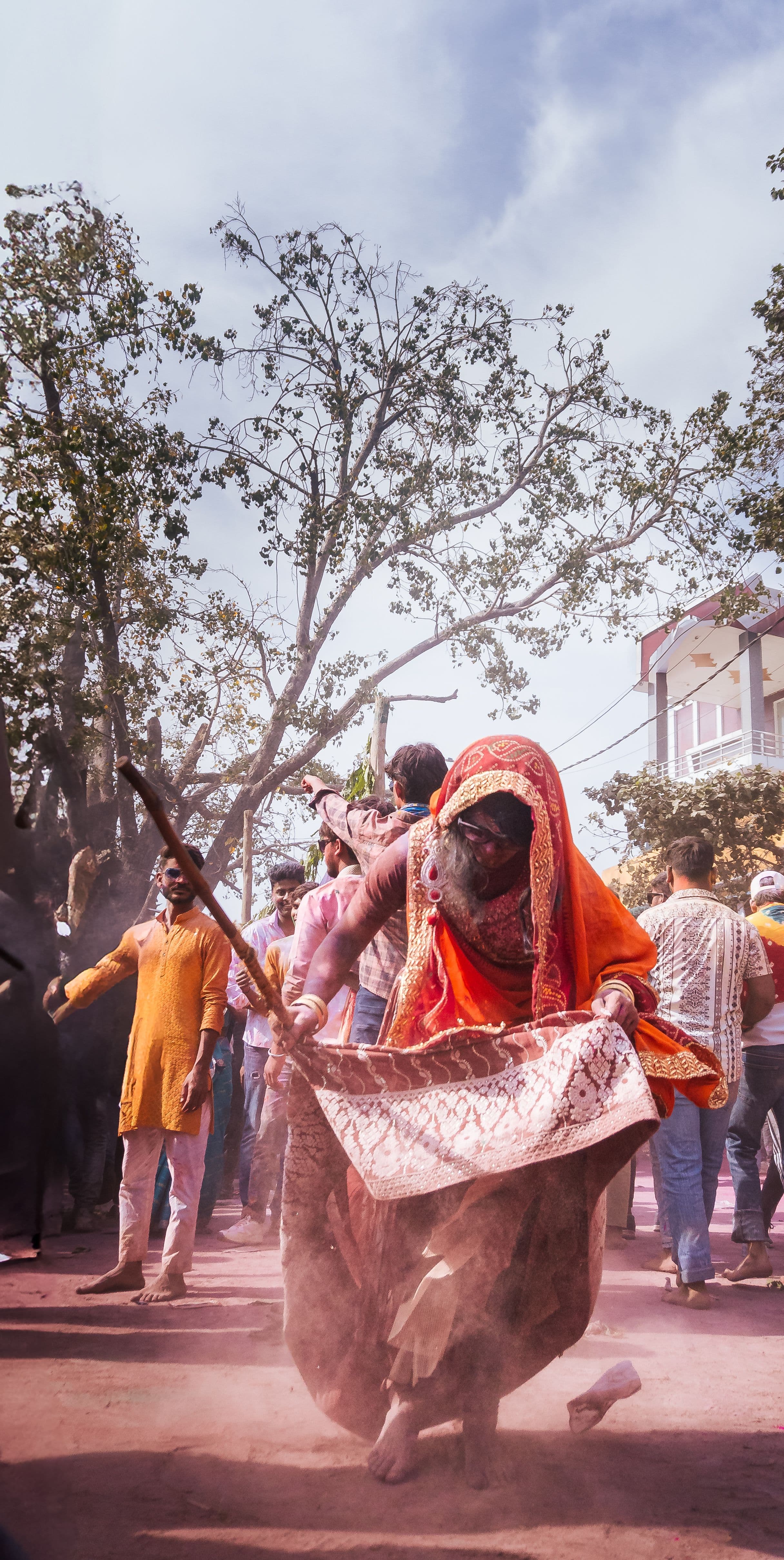 A woman in an orange sari raising a lath during Lathmar Holi, men shielding themselves in the lane behind