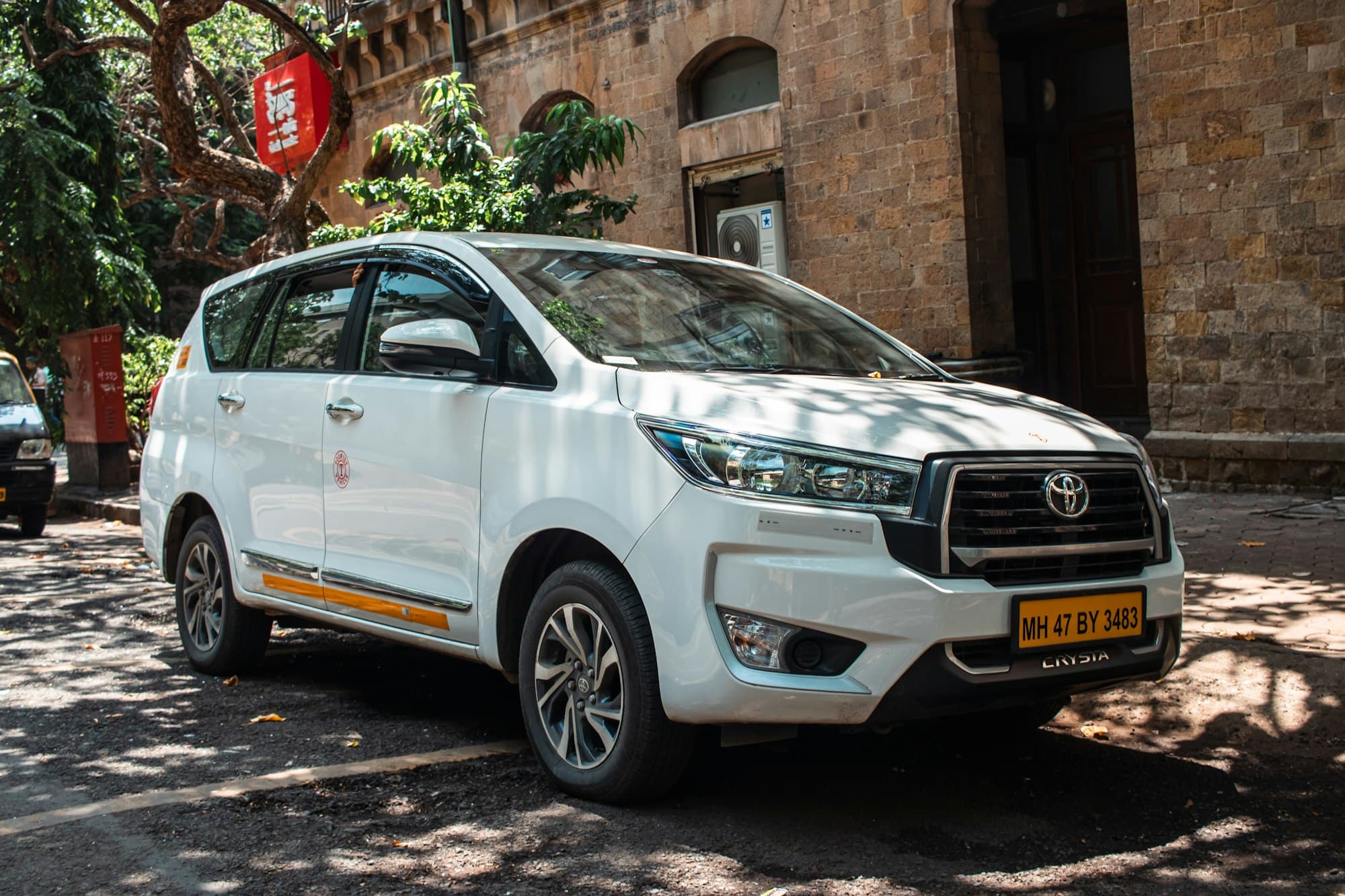 A white Toyota Innova Crysta in commercial taxi livery parked outside a stone building