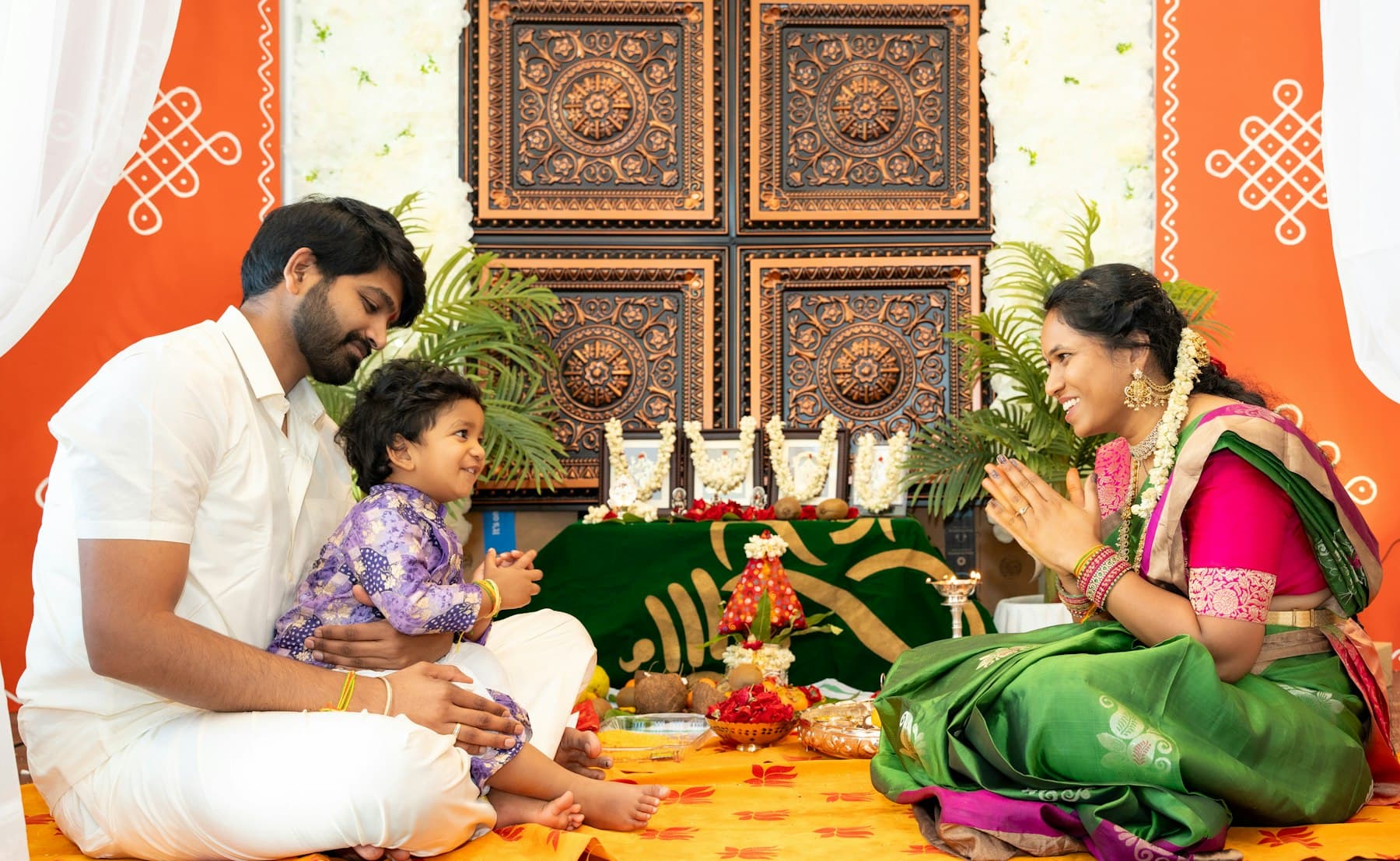 A family seated on the floor before a decorated shrine, the baby held between them