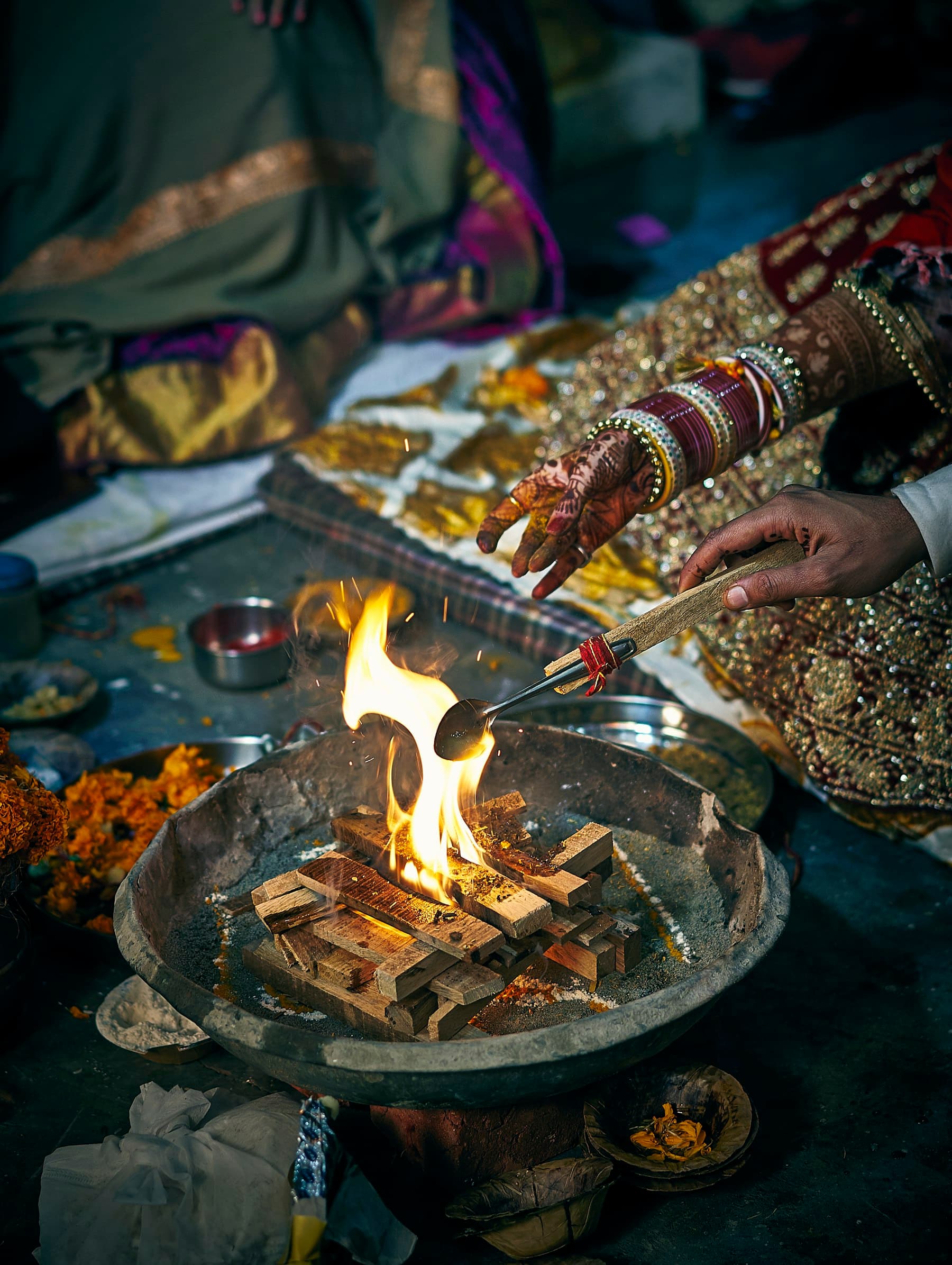 Hands feeding samagri into a small havan kund, the ingredients spread around it