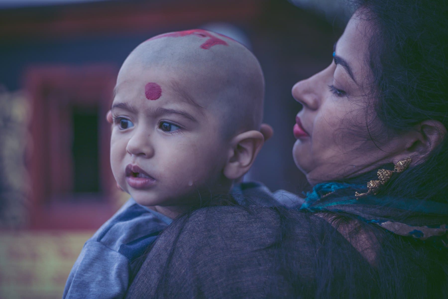 A small child with a freshly shaved head and a red tilak, held close by a woman